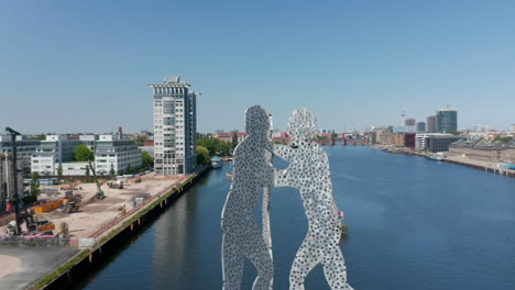 Rückwärts-Zeigen-Die-Molekülmann-Skulptur-In-Der-Spree-Und-Verschiedene-Gebäude,-Die-Ruhiges-Wasser-Umgeben.-Sonniger-Tag-Mit-Klarem-Himmel.-Berlin,-Deutschland