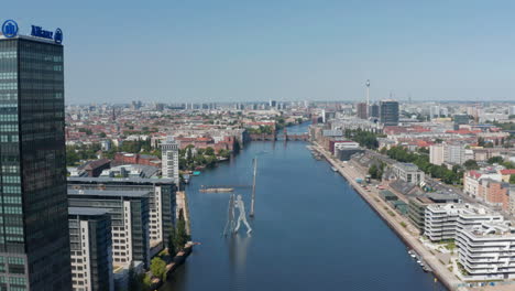 Forwards-descending-fly-above-Spree-river-towards-Molecule-Man-tall-sculpture.-Aerial-view-of-city.-Sunny-day-with-clear-sky.-Berlin,-Germany