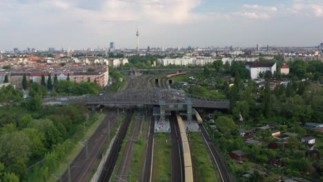 Forwards-fly-above-multi-track-railway-line-with-train-station.-Tracking-of-S-bahn-train-arriving-into-station-under-Bosebrucke-bridge.-Berlin,-Germany