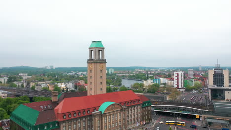 Tight-fly-around-town-hall-tower-with-tower-clock.-Historic-building-of-Rathaus-Spandau.-Revealing-panoramic-view-of-city-around-Havel-river.-Berlin,-Germany