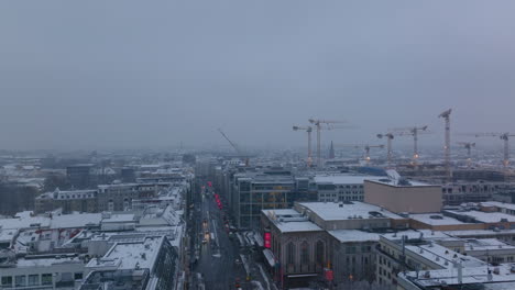 Aerial-view-of-snowy-buildings-in-large-town.-Group-of-tower-cranes-on-construction-site.-Cars-driving-on-street-at-dusk.-Berlin,-Germany