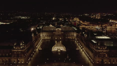 Aerial-view-of-large-glass-and-metal-pyramid-in-Louvre-Museum-complex.-Forwards-fly-above-buildings-at-night.-Paris,-France