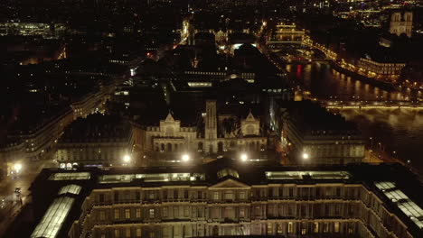 Forwards-fly-above-historic-buildings-of-Louvre-at-Seine-river-waterfront.-Illuminated-streets-in-night-city.-Paris,-France