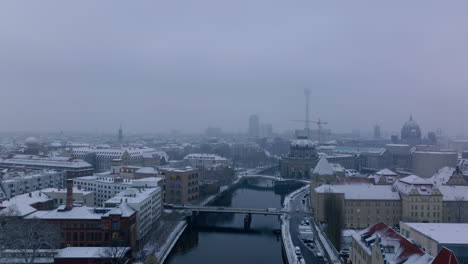 Backwards-fly-above-Spree-river-in-city-centre.-Cars-driving-on-bridge-across-water.-Snow-dusted-buildings-on-waterfront.-Berlin,-Germany