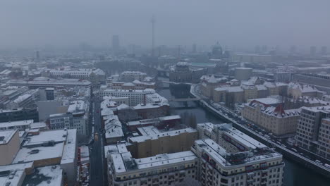 Forwards-fly-above-city-in-winter.-Various-buildings-around-river-with-snow-dusted-white-roofs.-Hazy-view-of-Berliner-dom-and-Fernsehturm.-Berlin,-Germany