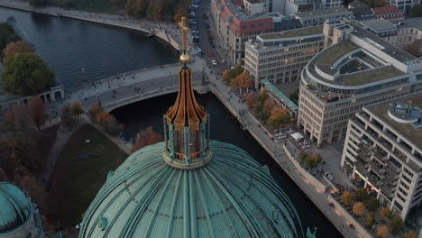 Orbit-shot-around-top-of-dome-with-religious-cross.-Aerial-view-of-riverbank-and-bridge-over-Spree-river-behind-roof-of-Berlin-Cathedral.-Berlin,-Germany.