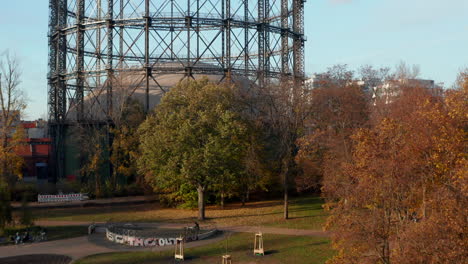 Famous-Gasometer-in-Berlin,-Germany-surrounded-by-Autumn-colored-Trees-and-Nature-in-Big-City,-Aerial-lift-up-revealing-shot