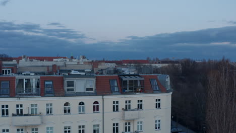 Aerial-slow-motion-view-of-traditional-brick-house-rooftop-across-street-with-vehicles-parked-and-moving-across-lane-surrounded-with-trees-on-a-cloudy-early-morning-in-Berlin,-Germany
