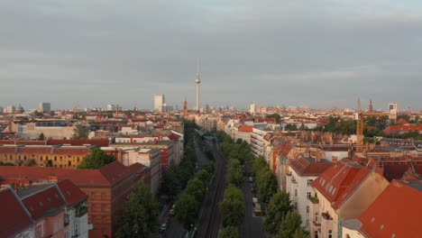 Aerial-view-of-morning-city-lit-by-bright-sunshine.-Fly-above-wide-street-with-railway-tracks-towards-Fernsehturm-TV-tower.-Berlin,-Germany