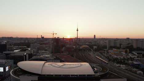 AERIAL:-Flight-over-Berlin,-Germany-at-beautiful-Sunset,-Sunlight-and-view-on-Alexanderplatz-TV-Tower-and-Ostbahnhof-and-Mercedes-Benz-Arena,-Sunflairs