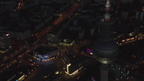 Primer-Plano-Del-Rascacielos-De-La-Torre-De-Televisión-De-Berlín-Por-La-Noche-Con-El-Paisaje-Urbano-Alemán-En-La-Distancia,-Toma-Aérea-De-Establecimiento