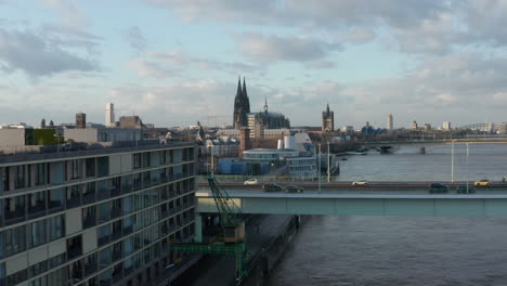 Forwards-fly-above-river-bank.-Busy-road-bridge-over-Rhine-river-and-Cologne-Cathedral-towering-high-above-surrounding-development.-Cologne,-Germany