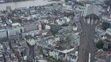 Forwards-fly-above-urban-neighbourhood.-High-angle-view-of-Main-train-station,-train-leaving-covered-platforms.-Cologne,-Germany