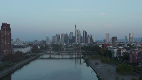 Empty-Frankfurt-am-Main-Skyline-in-early-morning-light-reflecting-in-Skyscrapers-with-Main-River-and-Bridges,-Aerial-sideways-slide-left