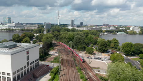 Aerial-view-of-train-unit-driving-on-multi-track-railway-line-leading-through-city.-Pleasant-environment-with-trees-and-water-area.-Free-and-Hanseatic-City-of-Hamburg,-Germany