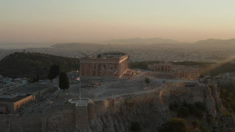 Aerial-Flight-towards-Acropolis-of-Athens-with-Greek-Flag-Waving-in-Beautiful-Golden-Hour-Sunset-light-with-Ocean-in-the-distance