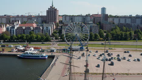 Backwards-reveal-of-waterfront-in-town.-Historic-sailboat-and-modern-sightseeing-boat-moored-on-shore.-Turning-Ferris-wheel-at-parking-lot.-City-in-background