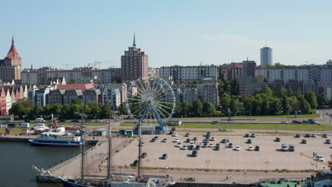 Slide-and-pan-shot-of-waterfront-with-moored-ships-and-Ferris-wheel-at-large-parking-lot.-Buildings-in-urban-neighbourhood-in-background