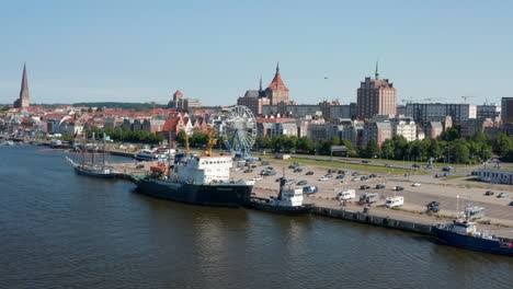 Fly-above-river-along-waterfront-with-parking-lot-with-Ferris-wheel-amusing-attraction.-Ships-moored-at-shore,-city-in-background