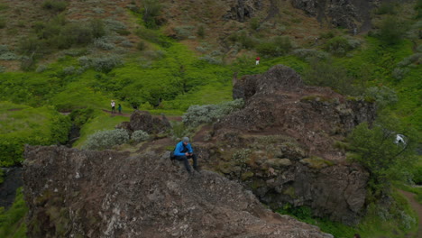 Aerial-view-man-trekker-sitting-peak-in-iceland-highlands-using-drone.-Birds-eye-view-man-tourist-on-top-peak-rock-formation-in-icelandic-countryside-driving-drone-with-pad