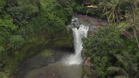 Aerial-shot-flying-towards-a-large-waterfall-in-the-Bali-jungle-with-outdoors-restaurant-on-the-cliff-above