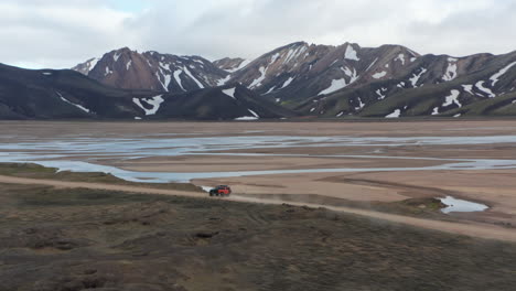 Vista-De-Drones-Increíble-Paisaje-De-La-Campiña-Islandesa-Con-Un-Alto-Pico-De-Montaña-Nevado.-Vista-De-Pájaro-Del-Vehículo-De-Expedición-Todoterreno-4x4-Acelerando-Conduciendo-Camino-De-Tierra-En-Las-Tierras-Altas-De-Islandia