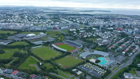 Aerial-footage-of-football-fields-and-swimming-pools.-Tilt-up-reveal-of-city-and-sea.-Overcast-sky.-Reykjavik,-Iceland