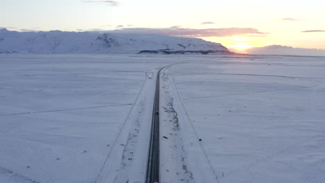 AERIAL:-Snow-White-Landscape-with-Road-following-Car-in-Iceland-Winter,-Sunset,-Arctic