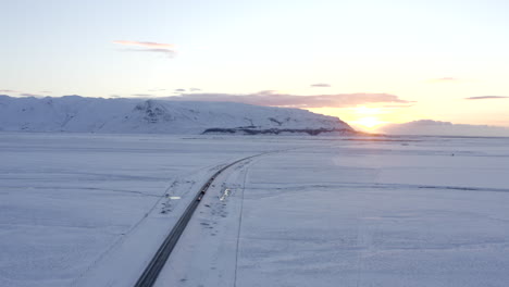 Antena:-Alto-Sobre-Paisaje-Blanco-Como-La-Nieve-Con-Carretera-Con-Coches-En-Islandia-Invierno,-Puesta-De-Sol,-ártico