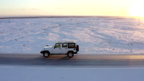 AERIAL:-Close-up-following-Jeep-from-side-on-snow-road-in-Iceland-at-Sunset-Winter,-Sun,-Arctic