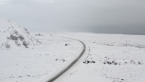 AERIAL:-Over-Snow-White-Landscape-with-Road,-Mountain-in-Iceland-Winter,-Snowing,-Cold,-Arctic