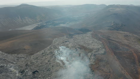 Forwards-fly-above-cooled-lava-stream-releasing-white-smoke.-Tilt-up-reveal-of-panoramic-landscape-view.-Fagradalsfjall-volcano.-Iceland,-2021