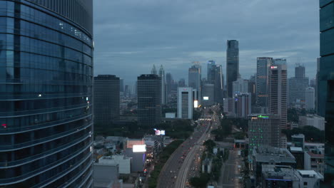 Aerial-rising-dolly-shot-between-two-skyscrapers-following-a-car-traffic-on-multi-lane-road-in-the-evening-in-Jakarta,-Indonesia