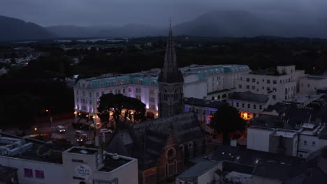 Pull-back-shot-of-historic-stone-church-with-tall-spire.-Backwards-fly-above-evening-town.-Killarney,-Ireland