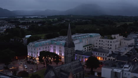 Historic-stone-church-with-tall-tower-and-luxury-hotel-building-with-illuminated-facade.-Traffic-in-evening-streets-of-town.-Killarney,-Ireland