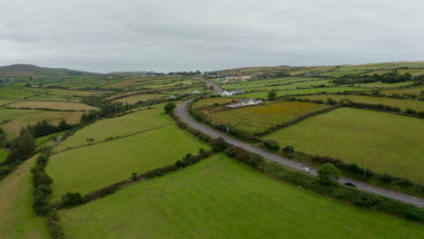Forwards-fly-above-cars-driving-on-road-between-green-meadows-and-pastures.-Cloudy-day-in-countryside.-Ireland