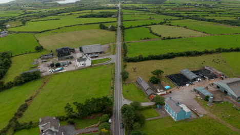 High-angle-view-of-road-leading-between-green-pastures-and-farmhouses.-Tilt-up-reveal-countryside-with-hills-in-background-and-cloudy-sky.-Ireland