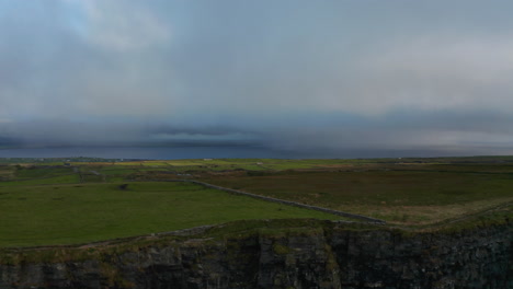 Rising-shot-of-rocky-cliff.-Revealing-green-meadows-and-pastures-and-sea-in-distance.-Overcast-sky.-Cliffs-of-Moher,-Ireland