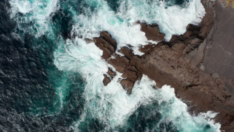 Aerial-birds-eye-overhead-top-down-view-of-mass-of-water-crashing-on-hard-stone-coast,-splashing-and-making-foam.-Kilkee-Cliff-Walk,-Ireland