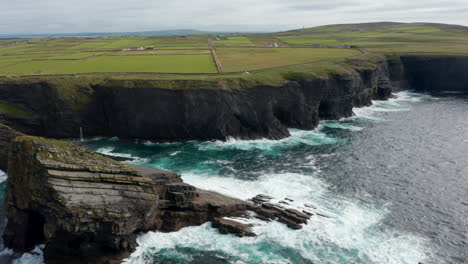 Aerial-ascending-footage-of-sea-coast-with-high-cliffs-and-broken-off-huge-stone-blocks.-Tilt-down-on-waves-washing-rocks.-Kilkee-Cliff-Walk,-Ireland