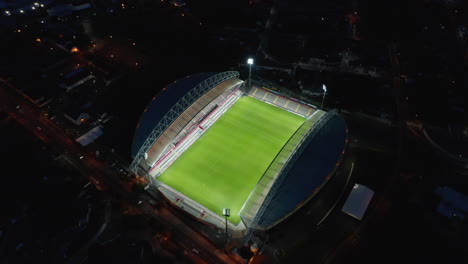 Fly-around-football-stadium-at-night.-High-angle-view-of-brightly-lit-empty-green-playfield,-sports-venue-in-city.-Limerick,-Ireland