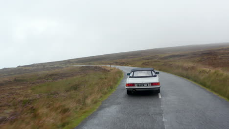 Forwards-tracking-of-silver-vintage-sports-car-driving-on-wet-road-along-moorlands.-Autumn-hazy-weather.-Ireland