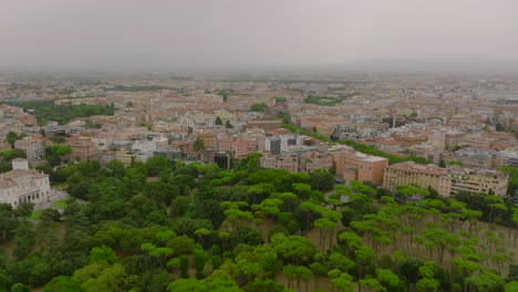 Green-trees-in-Villa-Borghese-park-and-apartment-buildings-in-urban-borough.-Forwards-fly-above-city-on-cloudy-day.-Rome,-Italy