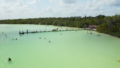 Fliegen-Sie-über-Holzpfeiler-Am-Seeufer-Mit-Menschen,-Die-Den-Heißen-Sonnigen-Tag-Genießen.-Naturschwimmbad-Mit-Seichtem-Wasser-In-Tropischer-Destination.-Kaan-Luum-Lagune,-Tulum,-Yucatan,-Mexiko