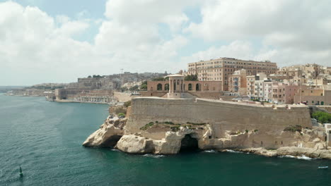 Famous-Siege-Bell-War-Memorial-in-Valletta,-Malta-capital-City,-Aerial-Drone-backwards,-Revealing-a-passing-Cargo-Ship