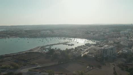 Bahía-Con-Barcos-Y-Sol-Reflejados-En-El-Agua-Al-Atardecer-En-La-Isla-Mediterránea-De-Malta,-Vista-Aérea-De-Drones
