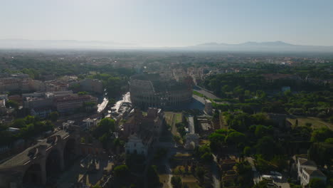 Aerial-slide-and-pan-footage-of-monumental-ancient-Colosseum-amphitheatre-and-surrounding-buildings-and-park-in-city.-Rome,-Italy