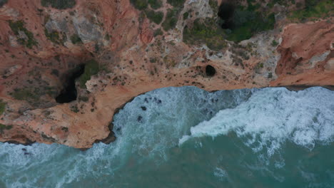 Sensational-top-down-drone-aerial-view-of-turquoise-water-waves-crashing-against-cliff-coast,-Lagos,-Portugal,-day