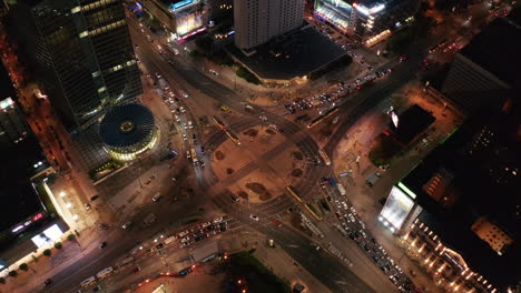 High-angle-view-of-large-and-busy-road-intersection-in-city-at-night.-Two-tram-units-passing-by-each-other-in-middle-of-roundabout.-Warsaw,-Poland