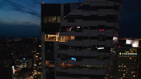 Fly-around-modern-futuristic-skyscraper-with-glossy-glass-facade.-Panoramic-view-of-night-cityscape.-Warsaw,-Poland